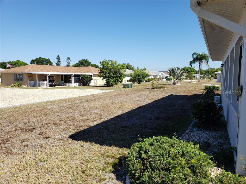 Side yard beside a home, with dry grass, shrubs, and neighboring houses across the street.