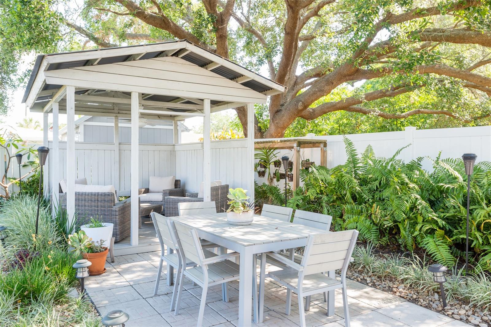 Outdoor dining patio with a white pergola, wicker lounge seating, and lush landscaping beneath a large tree.