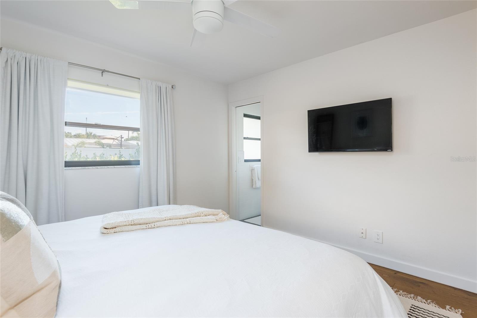 Bedroom with a white bed, window, wall-mounted television, and open bathroom doorway.