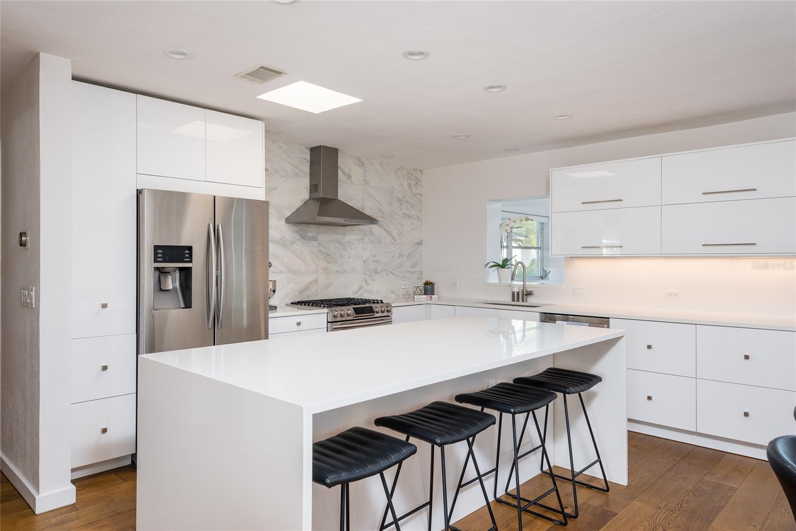 Modern white kitchen with a large island, black bar stools, stainless steel appliances, and marble-pattern backsplash.