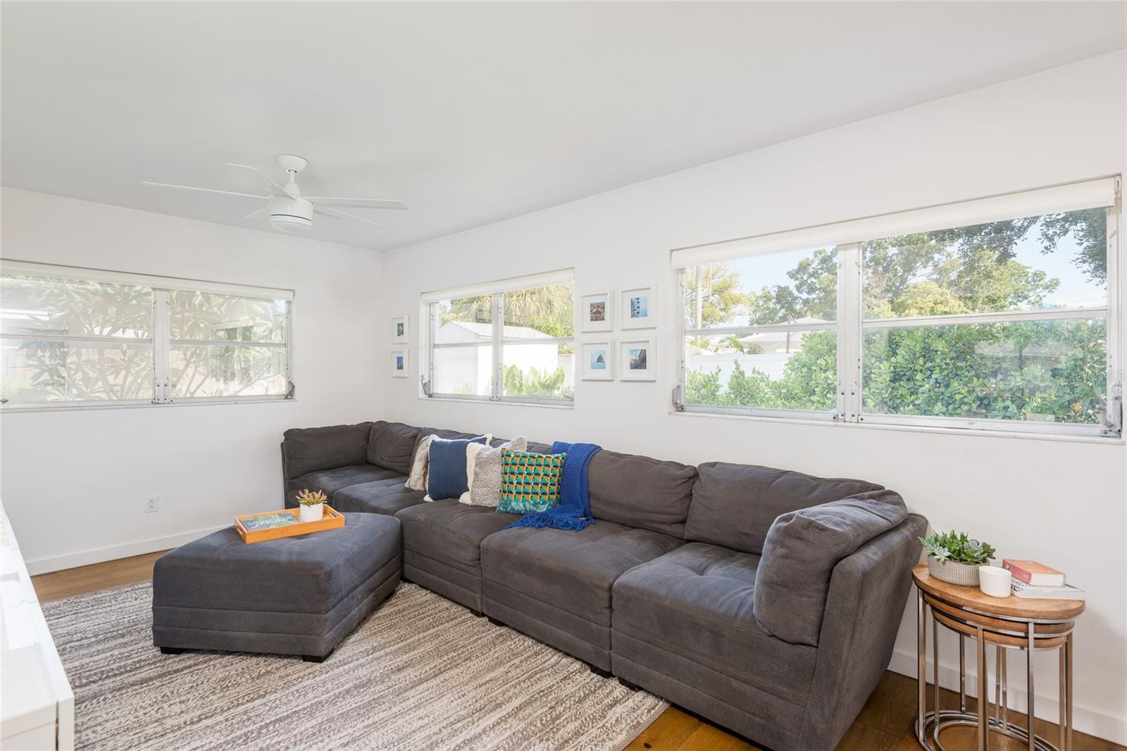 Bright living room with a gray sectional and chaise beneath wide windows overlooking greenery.