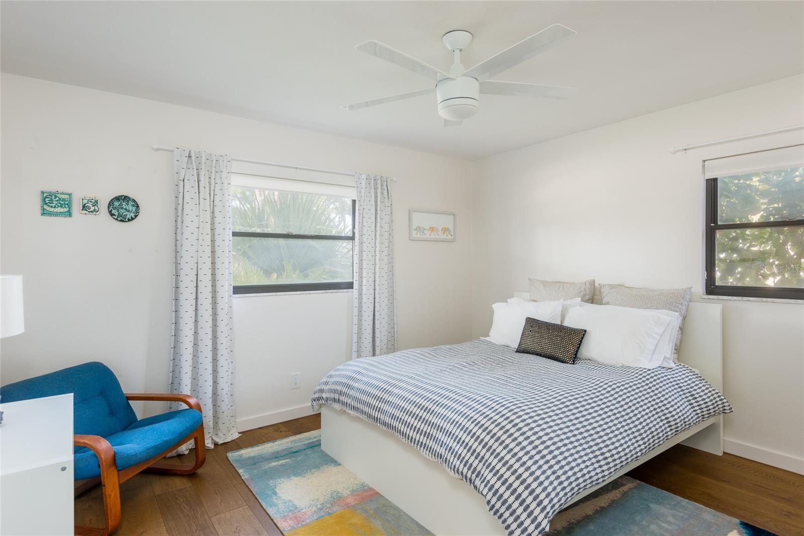 Furnished bedroom with patterned bedding, a blue chair, two windows, and a white ceiling fan.