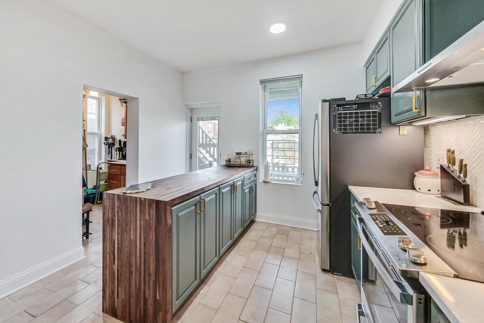 Kitchen with a long butcher-block island, green-gray cabinets, and stainless steel appliances.