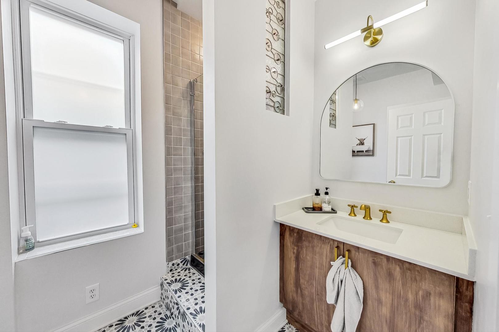 Bathroom with a wood vanity, brass fixtures, rounded mirror, and patterned tile shower floor.
