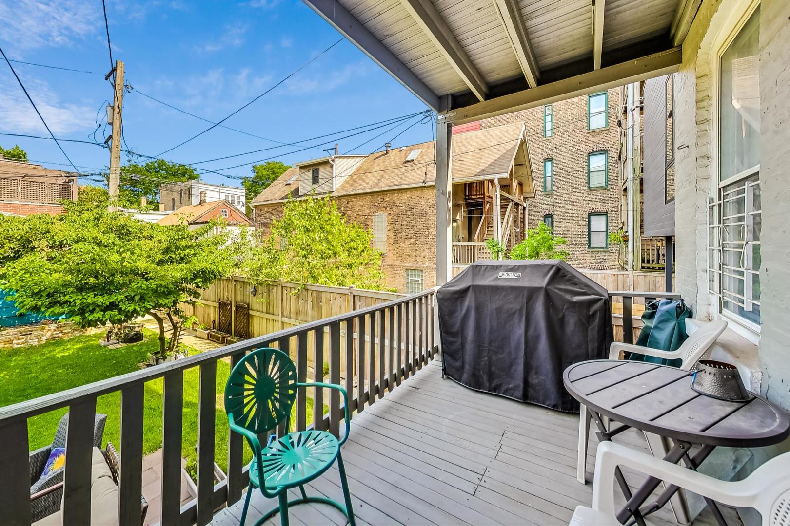Covered deck with a grill and seating overlooks a fenced backyard and leafy tree.