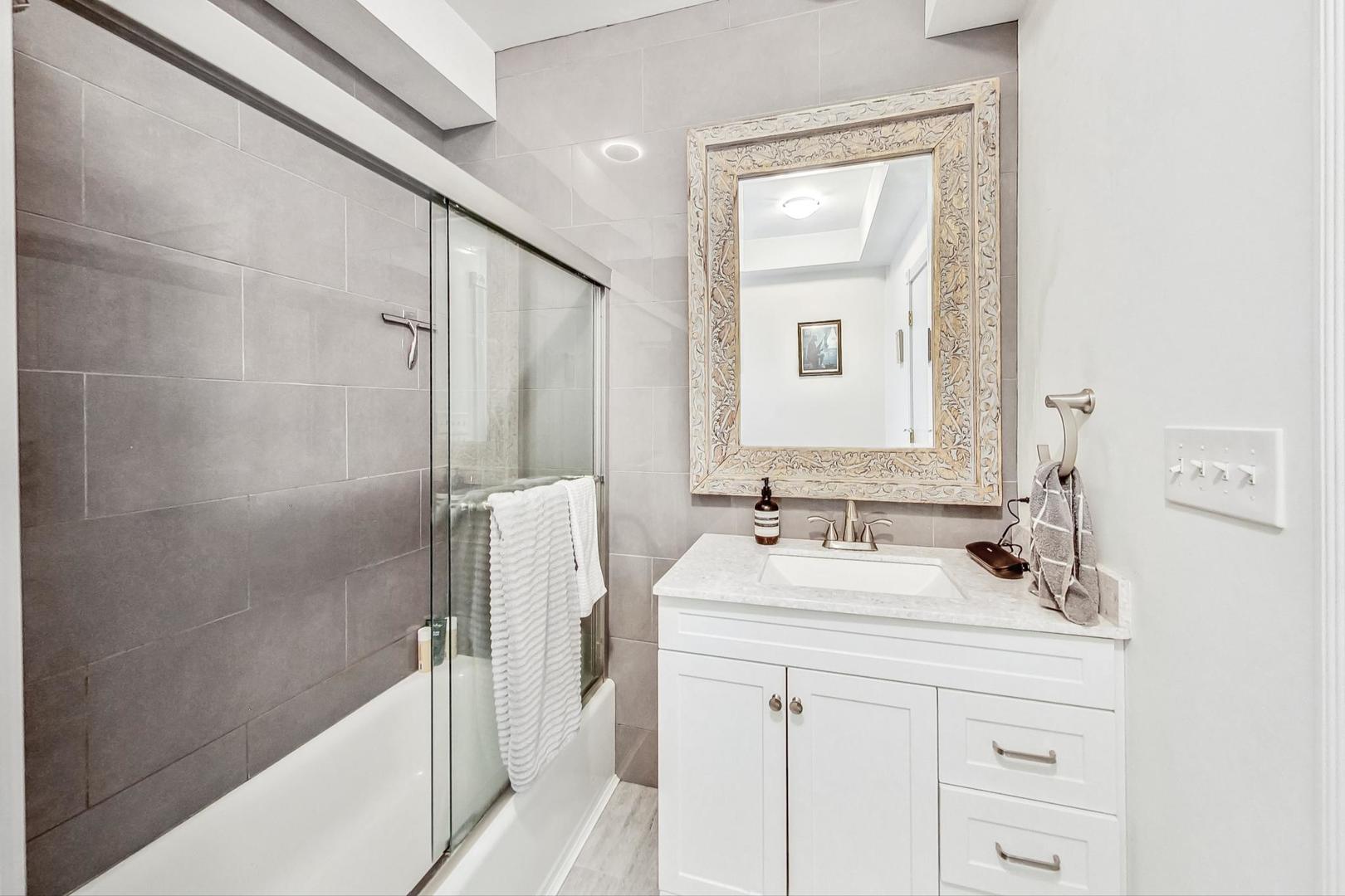 Bathroom with a glass-enclosed tub, gray tile walls, and a white vanity with an ornate mirror.