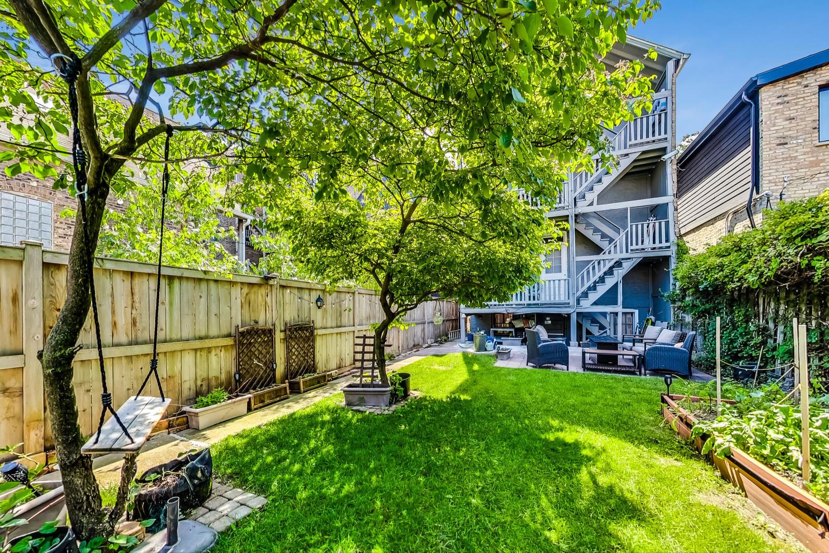 Fenced backyard with a sunny lawn, leafy trees, raised garden beds, and a furnished patio.