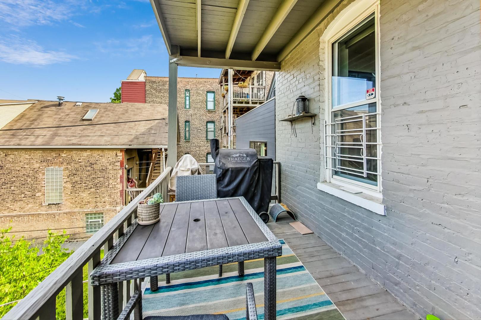 Covered balcony with a wicker dining table, striped rug, and grill beside a gray brick wall.