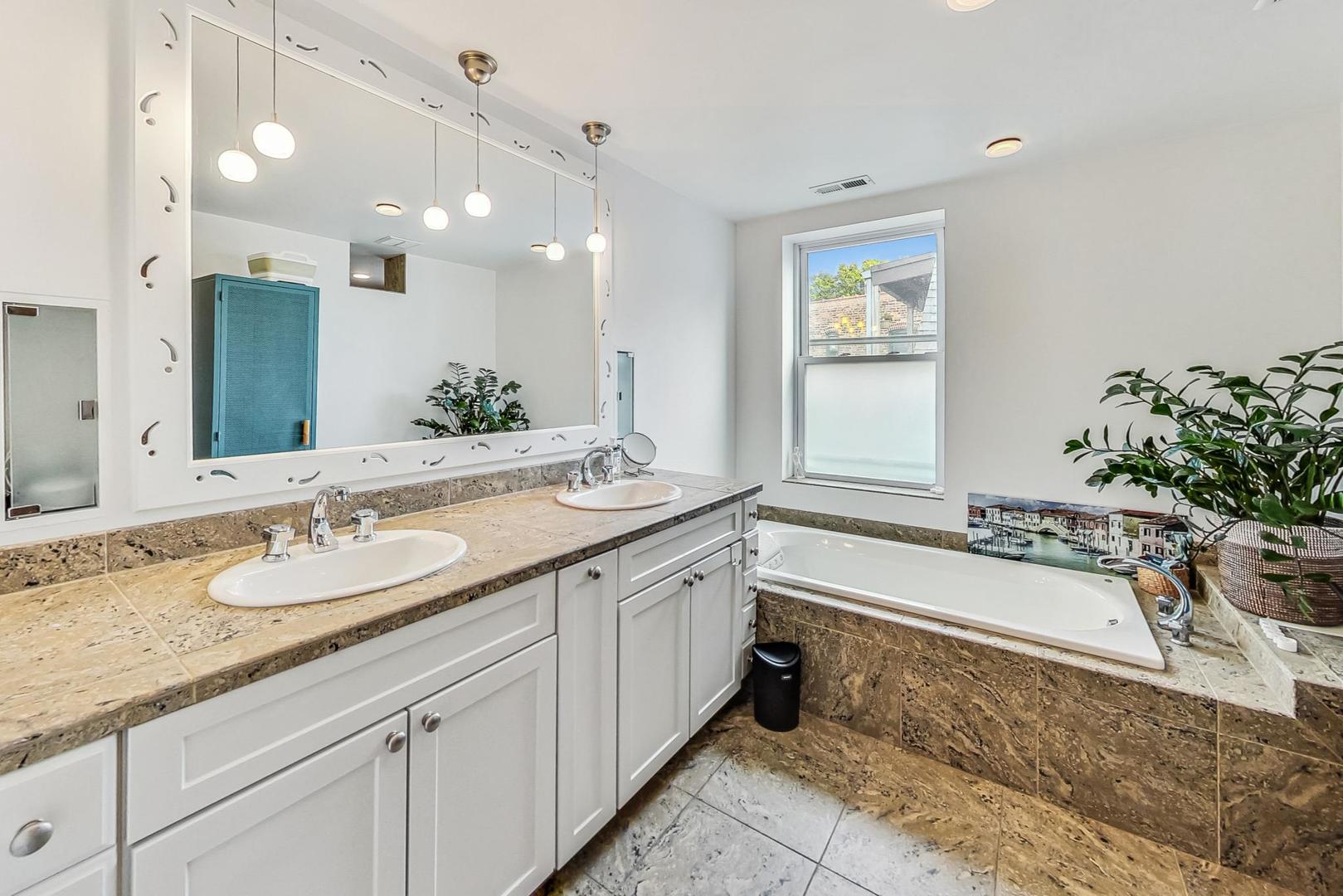 Bathroom with a double-sink vanity, large mirror, stone tile, and built-in soaking tub.