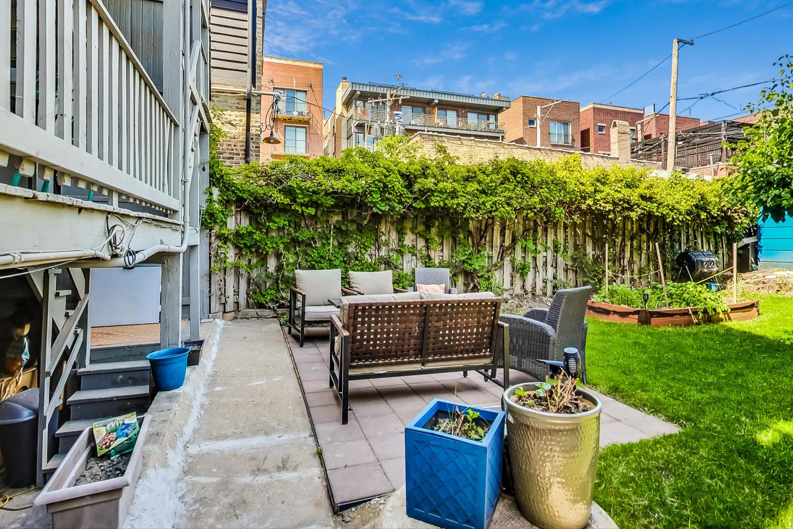 Backyard patio with outdoor seating beside a lawn, raised garden beds, and a vine-covered fence.