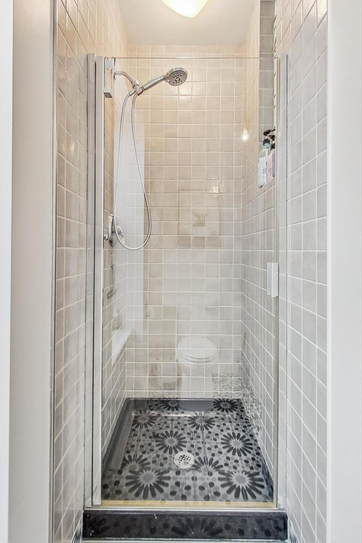 Walk-in shower with glass doors, pale tile walls, and a dark floral-patterned floor.