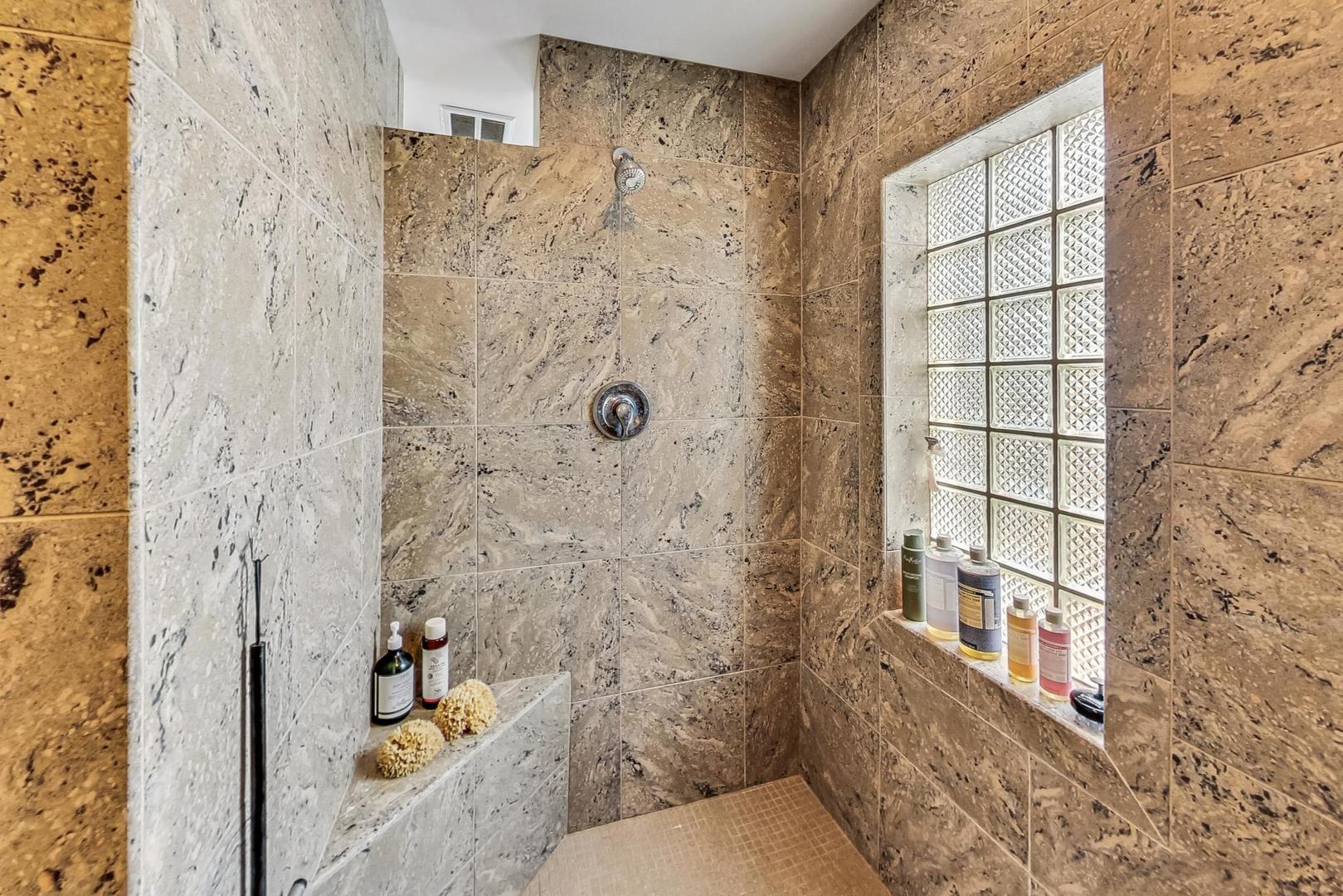Stone-tiled walk-in shower with a glass-block window and built-in ledge.