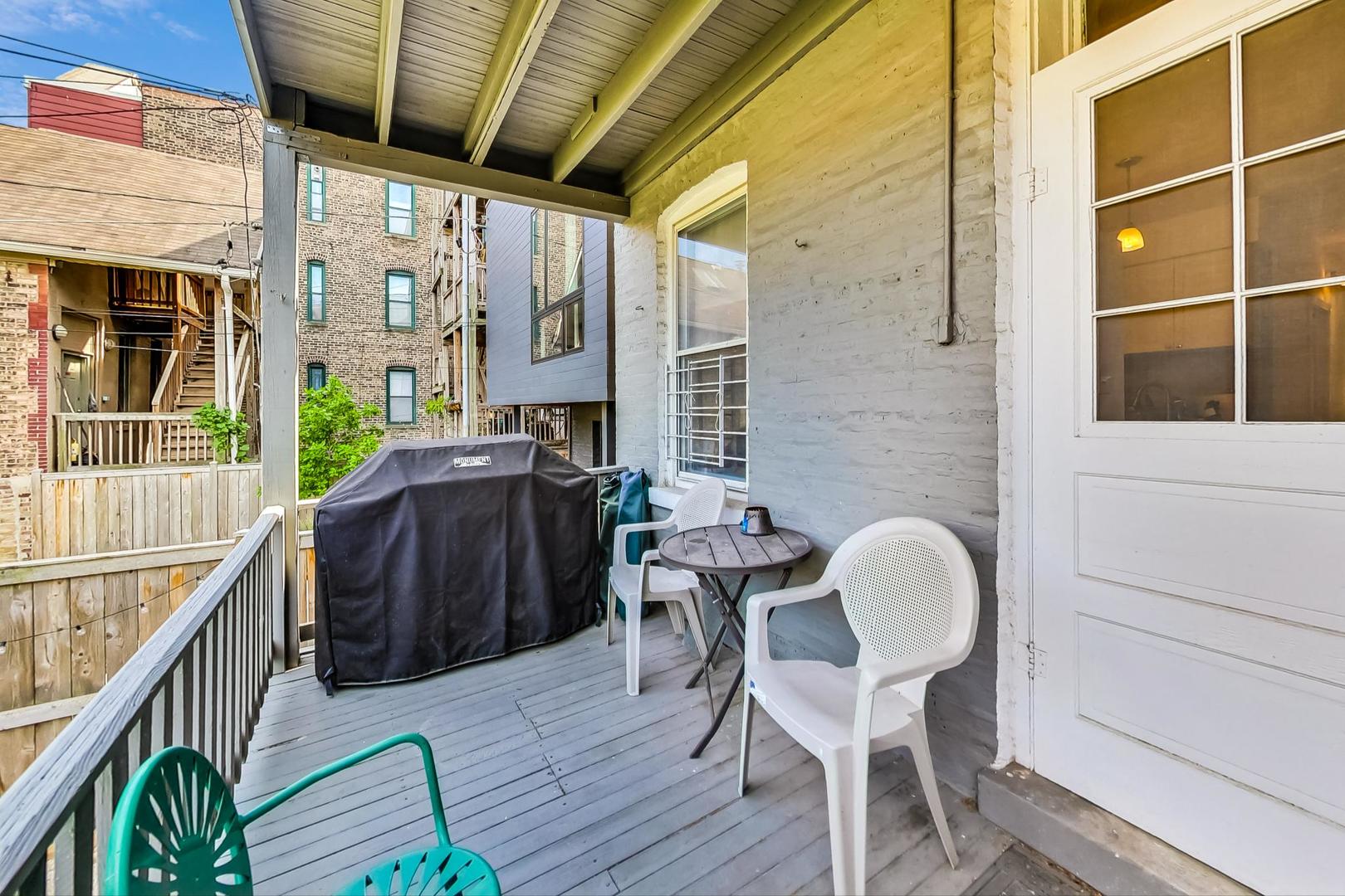 Covered porch with white chairs, a small table, and a covered grill beside a white brick wall.