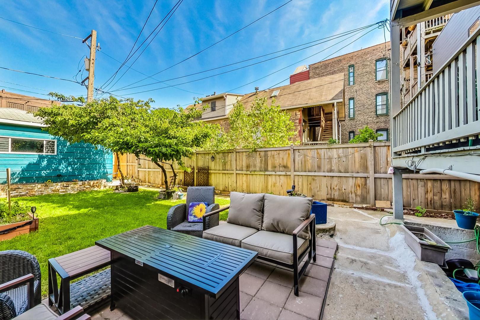 Backyard patio with lounge seating and table beside a leafy tree and lawn.