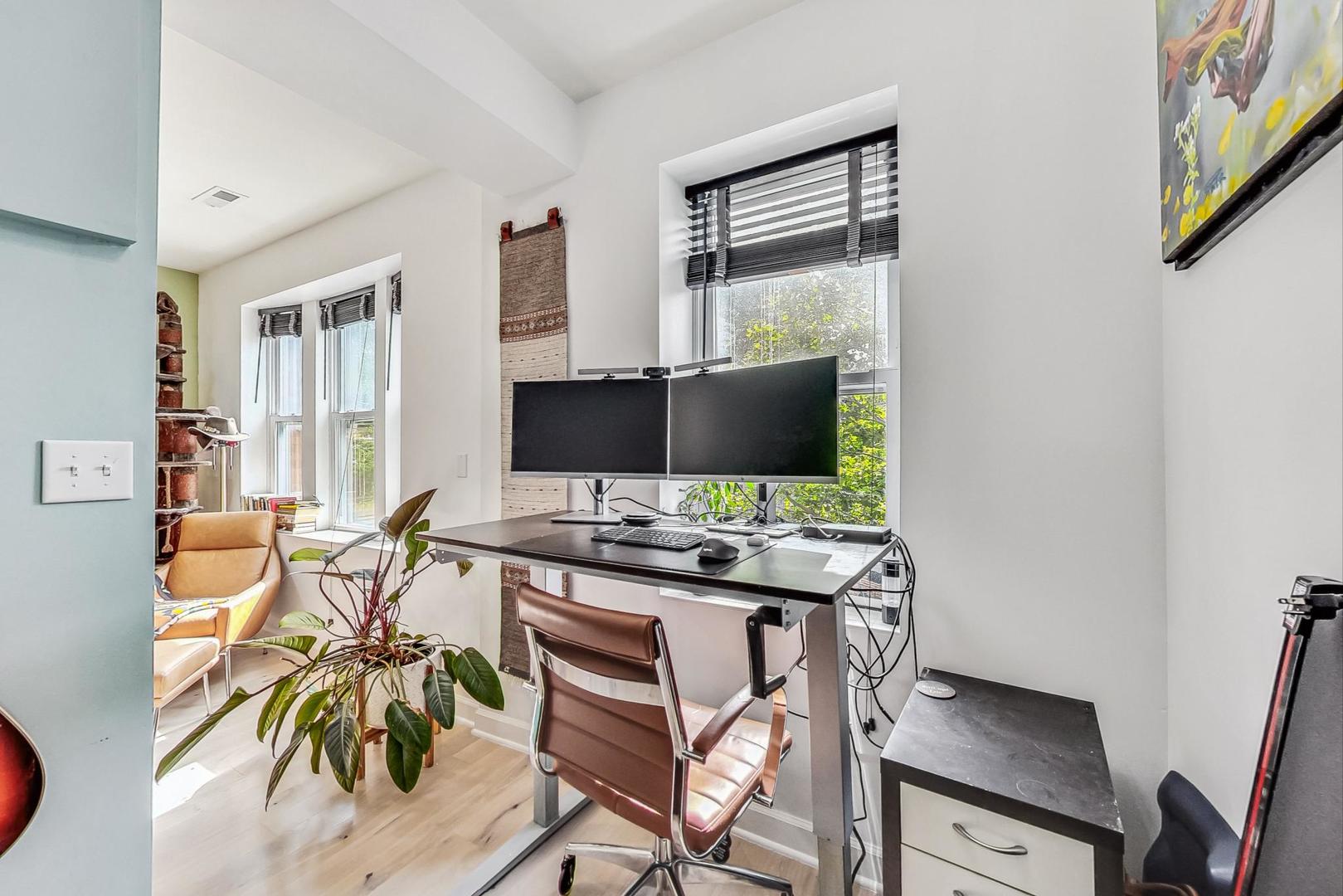 Home office with a standing desk, dual monitors, and a leafy plant beside tall windows.
