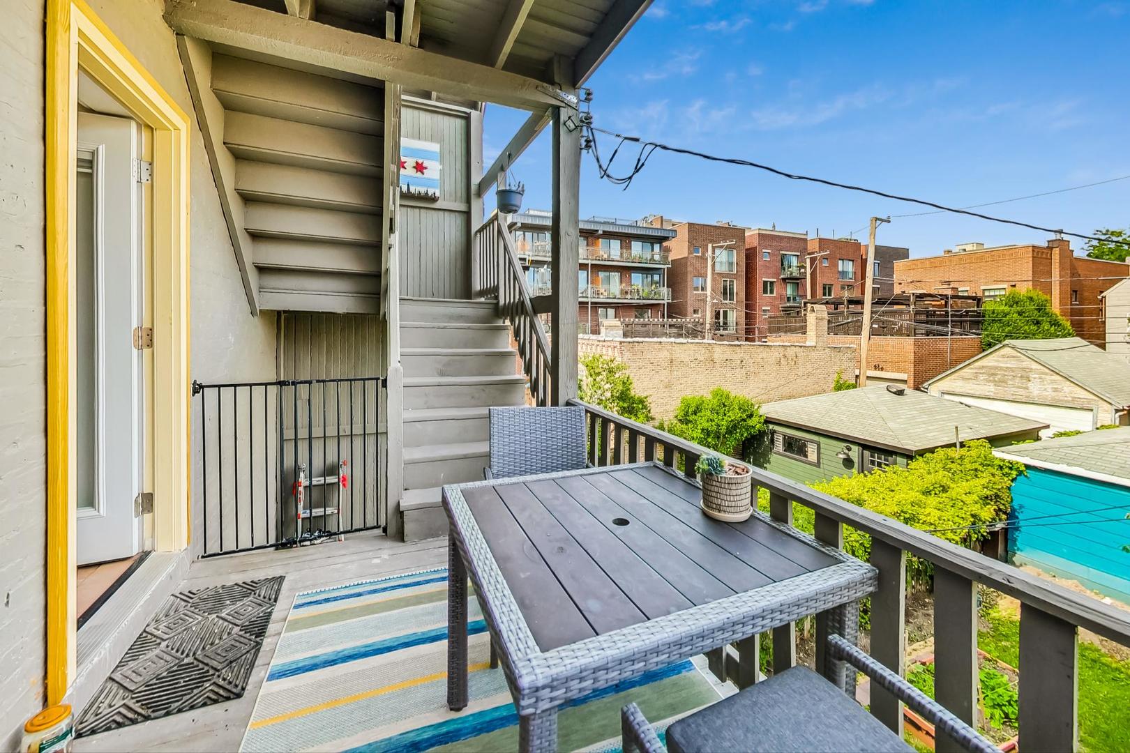 Furnished elevated deck with outdoor dining table, stairs, and views of neighboring buildings.