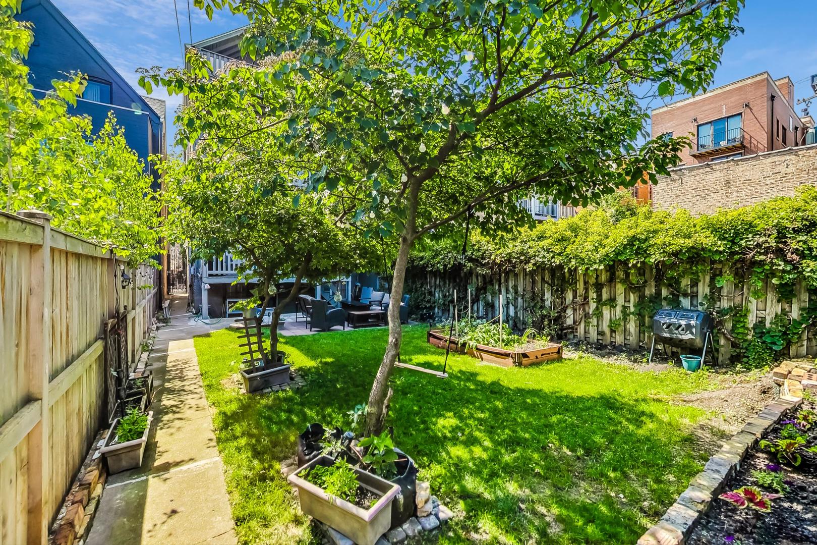 Backyard with a shaded lawn, patio seating, raised garden beds, and a paved path.