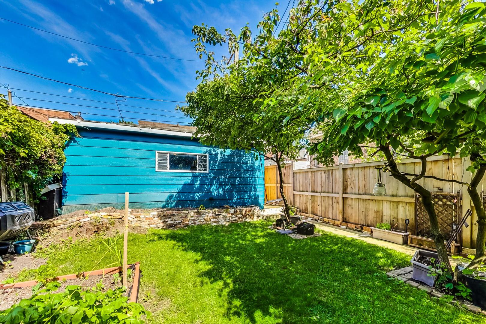 Backyard with a green lawn, mature shade trees, raised garden beds, and a bright blue detached garage.