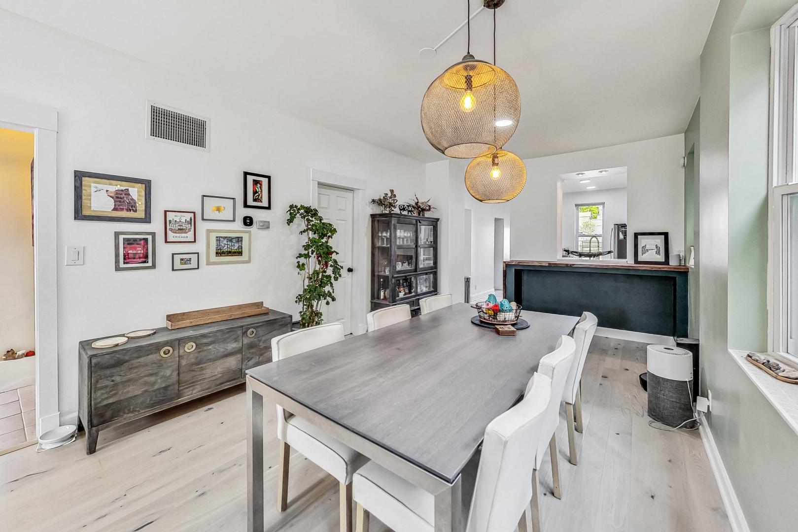 Dining room with a long gray table, white chairs, woven pendant lights, and a dark breakfast bar.