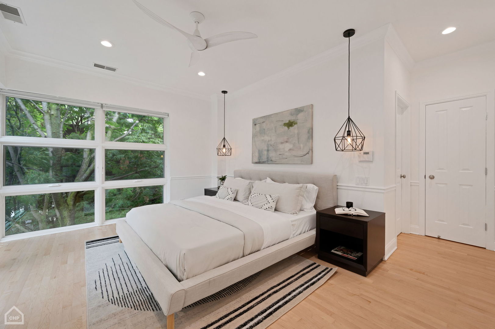 Staged bedroom with a large bed beside floor-to-ceiling windows overlooking leafy trees.