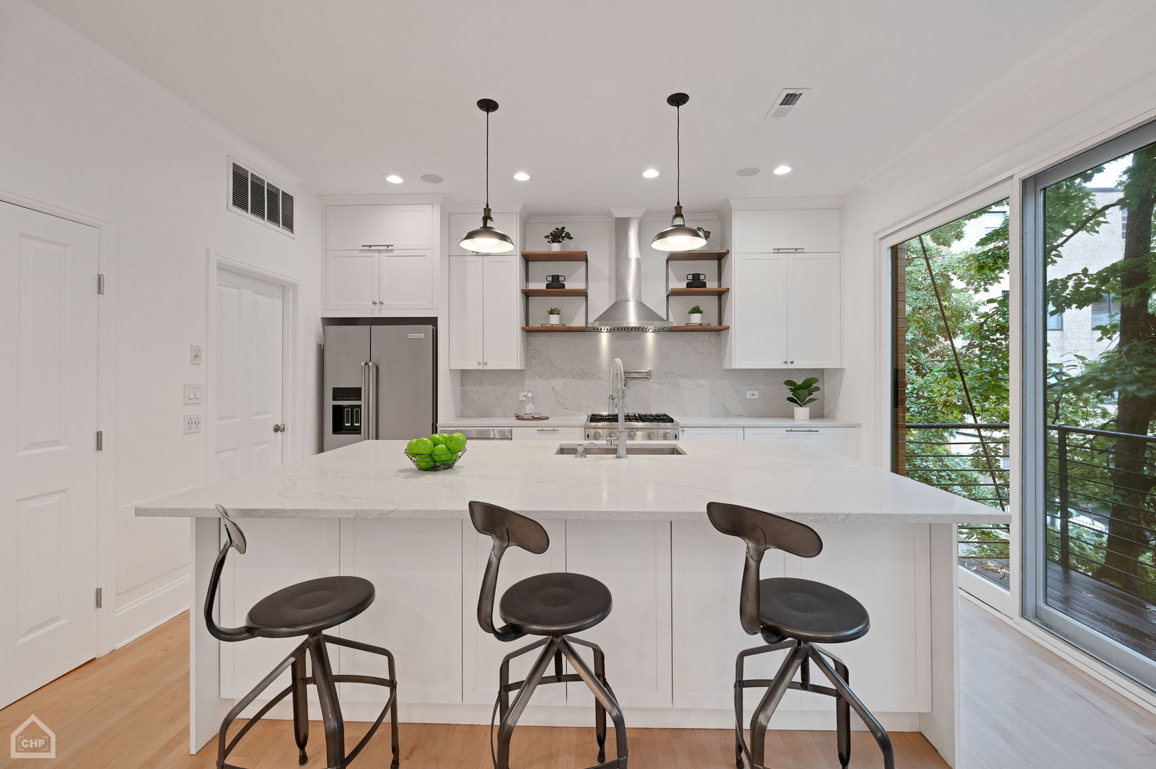 Bright white kitchen with a large stone island, three dark stools, and glass doors overlooking greenery.