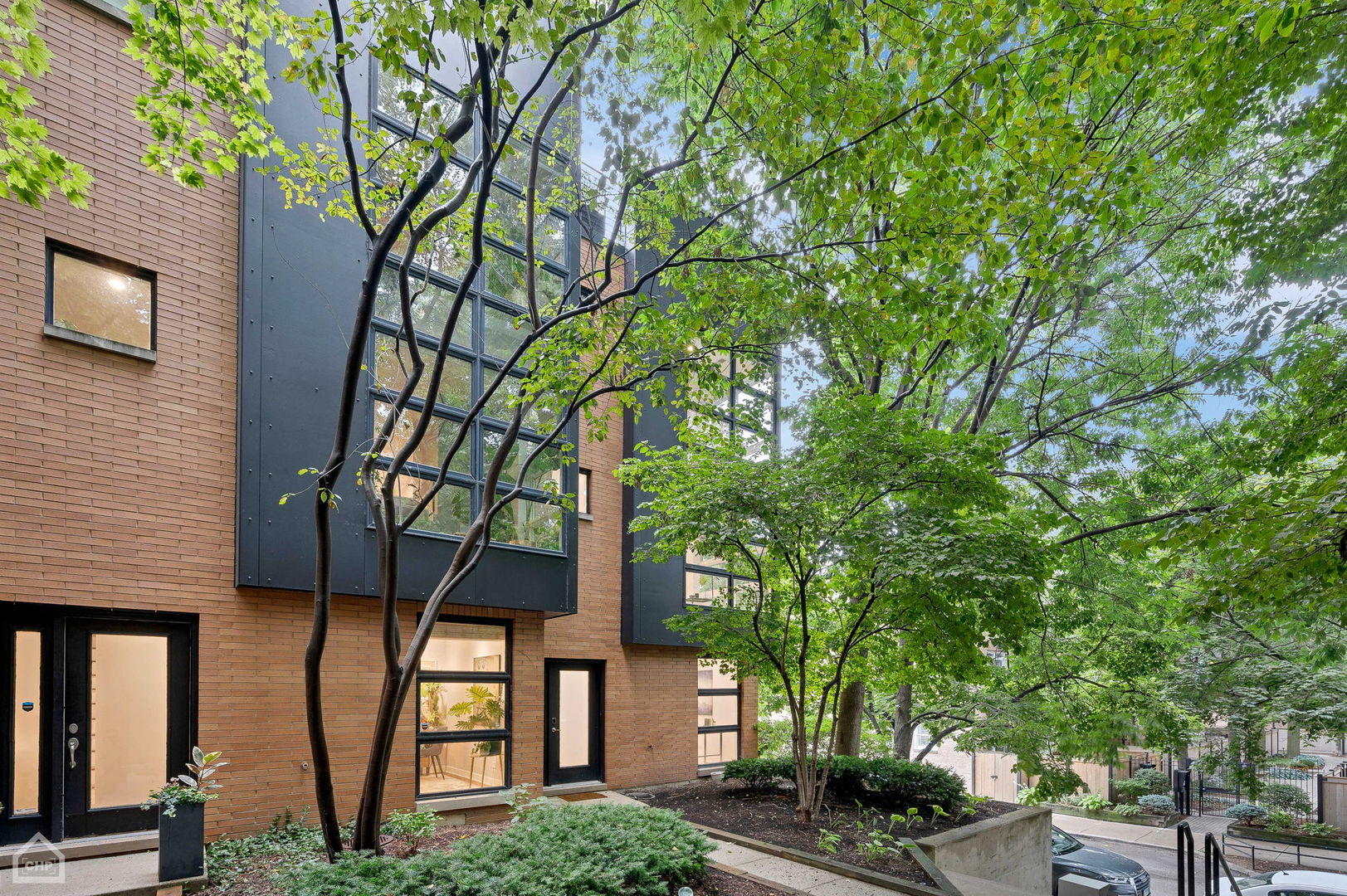 Modern brick residence with dark-framed windows, mature trees, and landscaped planting beds.