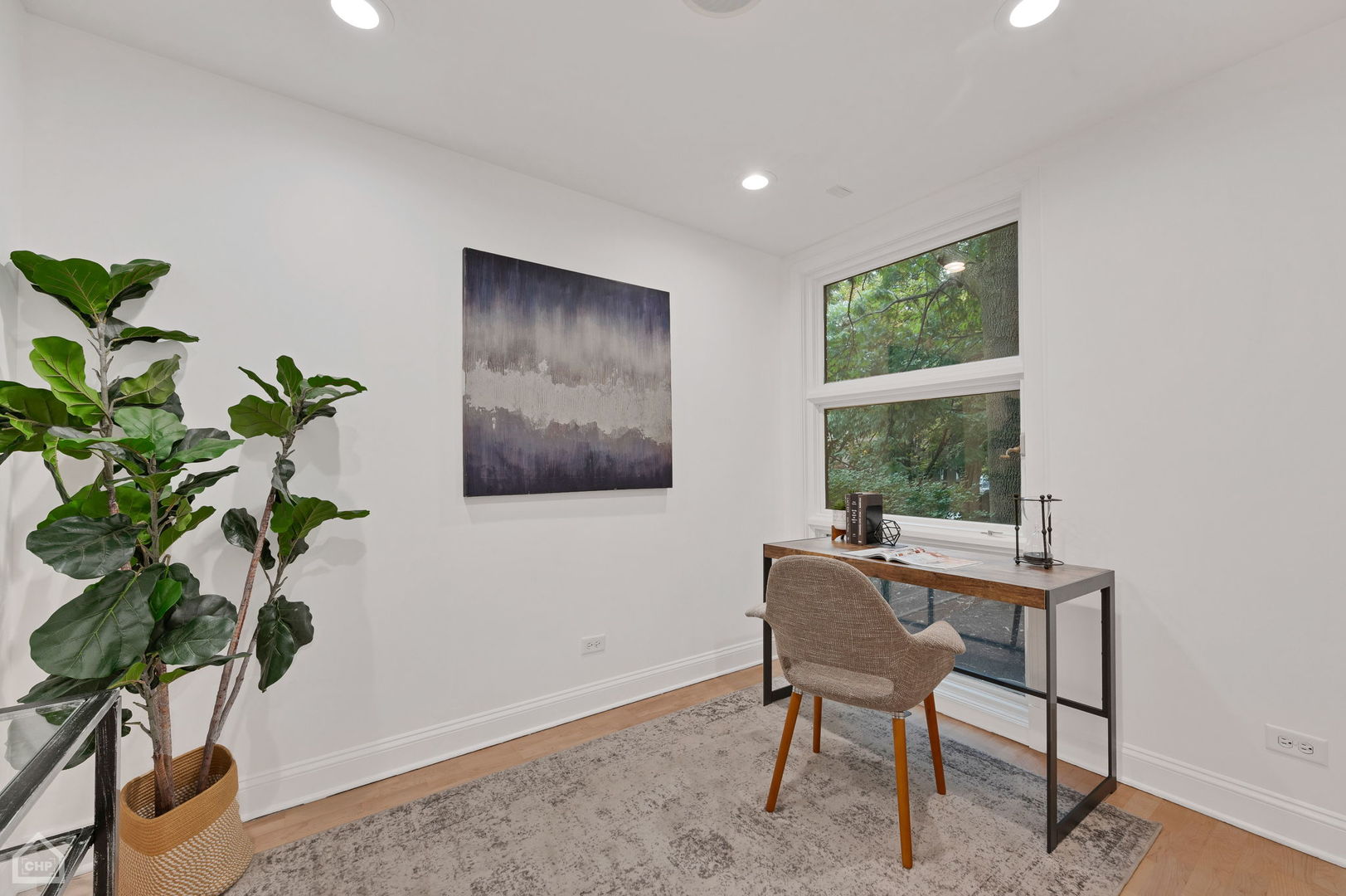 Furnished home office with a desk beside a large window overlooking leafy trees.