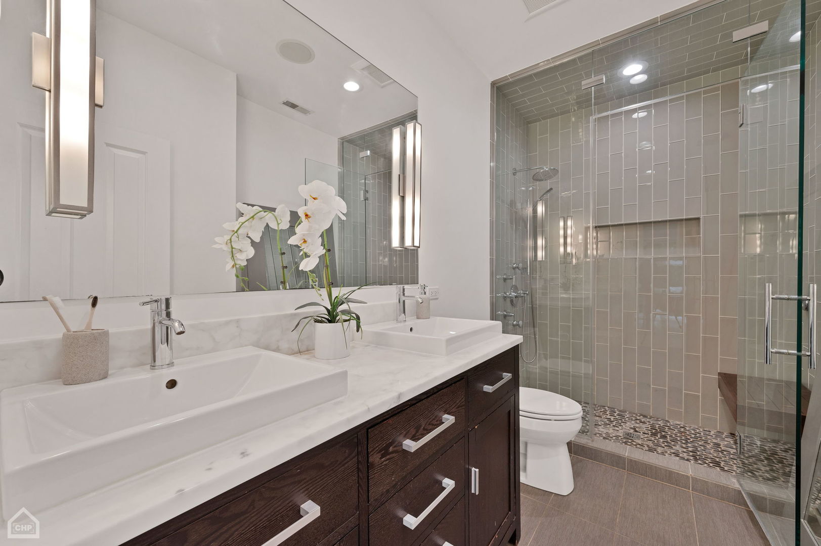 Bathroom with a double-sink vanity and glass shower tiled in gray, with pebble flooring.