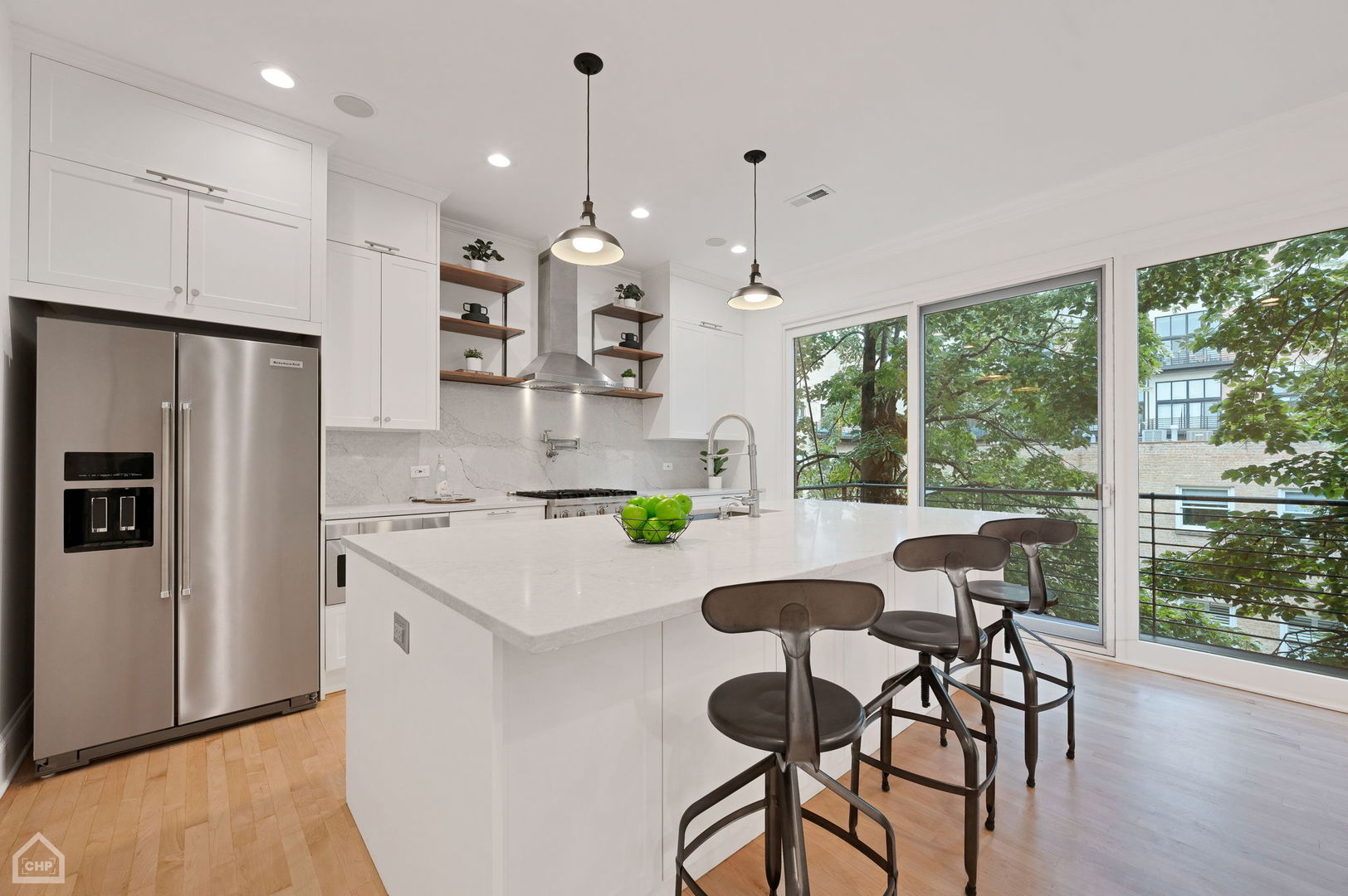 Modern white kitchen with a large island, bar stools, and wide windows overlooking leafy trees.