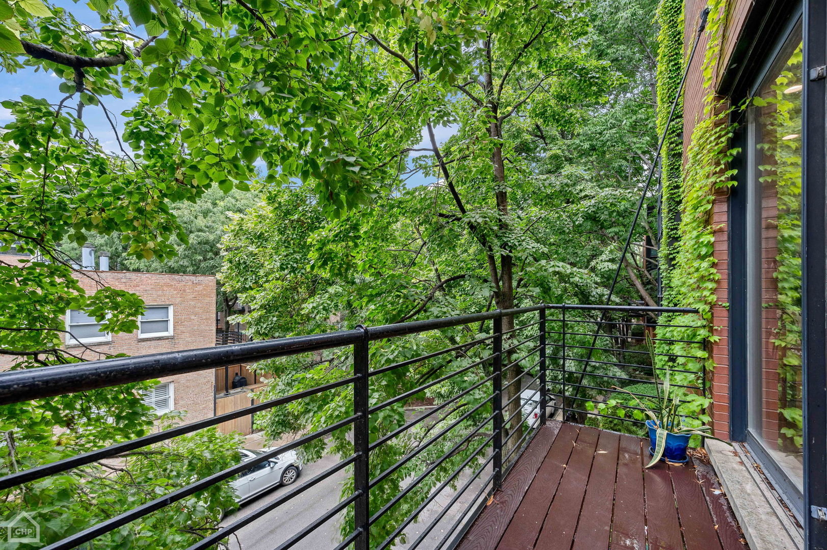 Wood-plank balcony surrounded by leafy trees, with black metal railings and a potted plant.