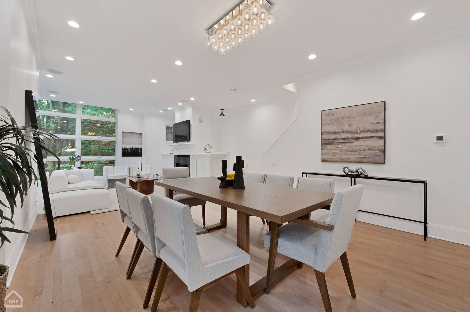 Open-plan dining and living room with a wooden table, fireplace, and large windows overlooking greenery.