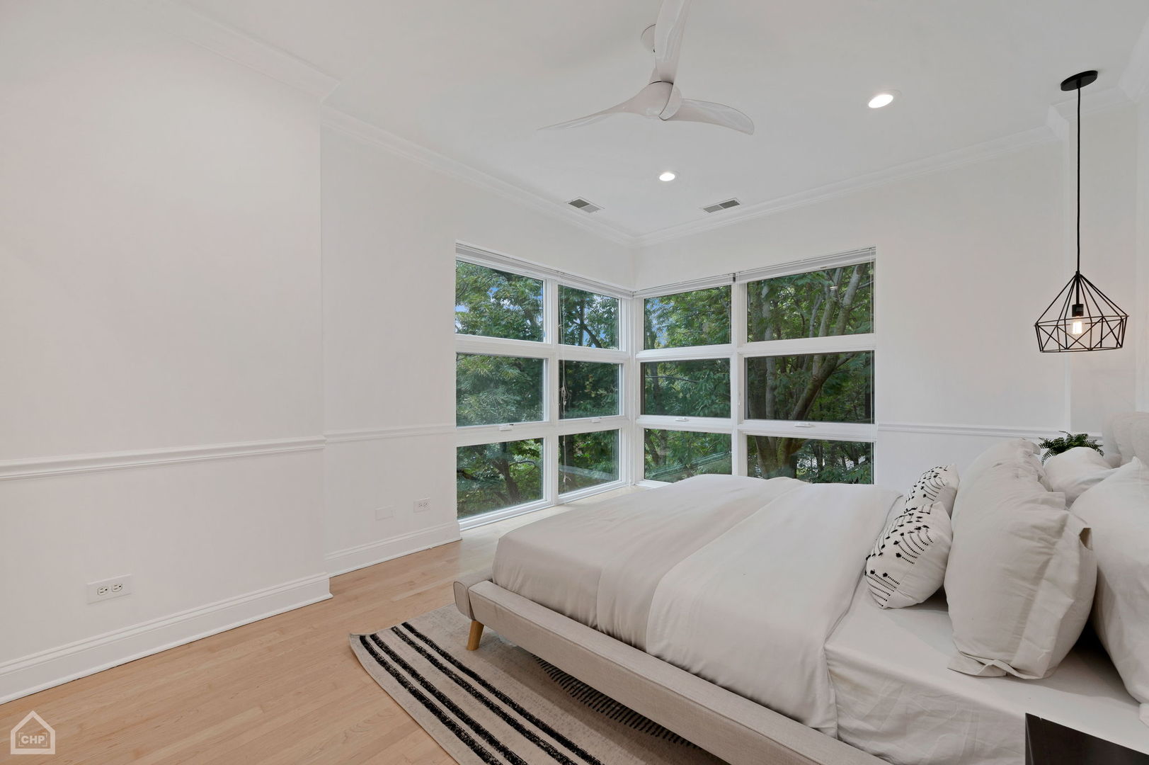 Bright bedroom with a white bed and large corner windows overlooking leafy trees.