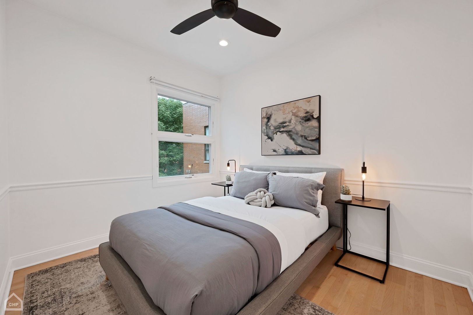 Staged bedroom with a gray bed, large window, black ceiling fan, and hardwood flooring.