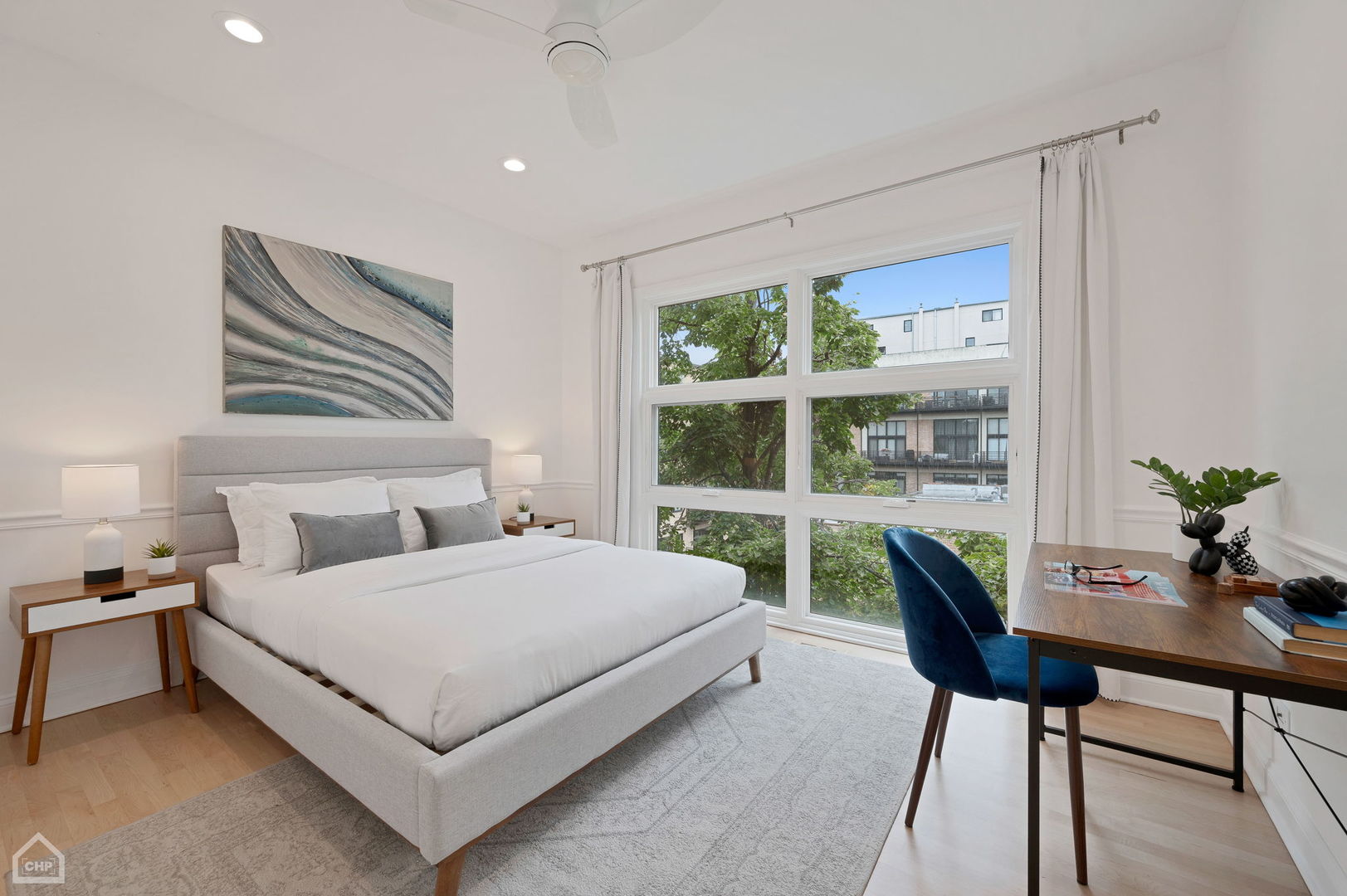 Staged bedroom with a gray upholstered bed, large window, and desk with a blue chair.
