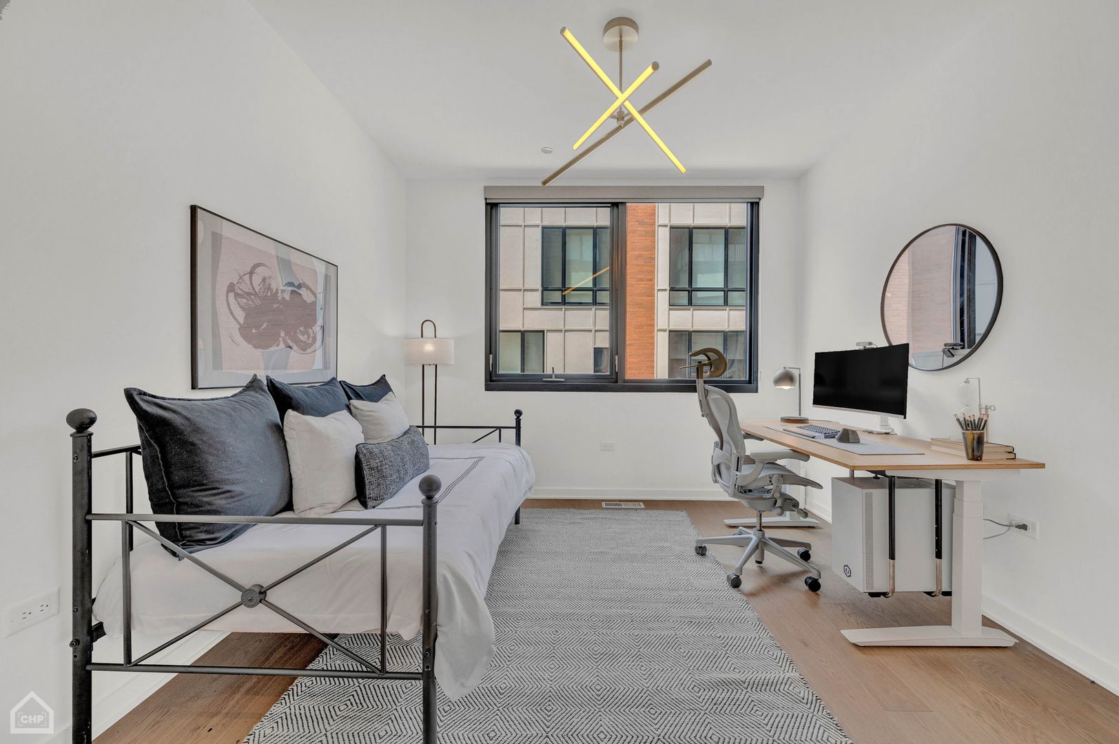 Furnished bedroom with a metal daybed, geometric rug, and desk beneath a large window.