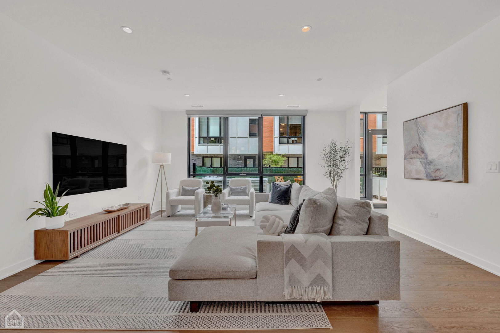 Staged condo living room with a gray sectional, white armchairs, and floor-to-ceiling courtyard windows.