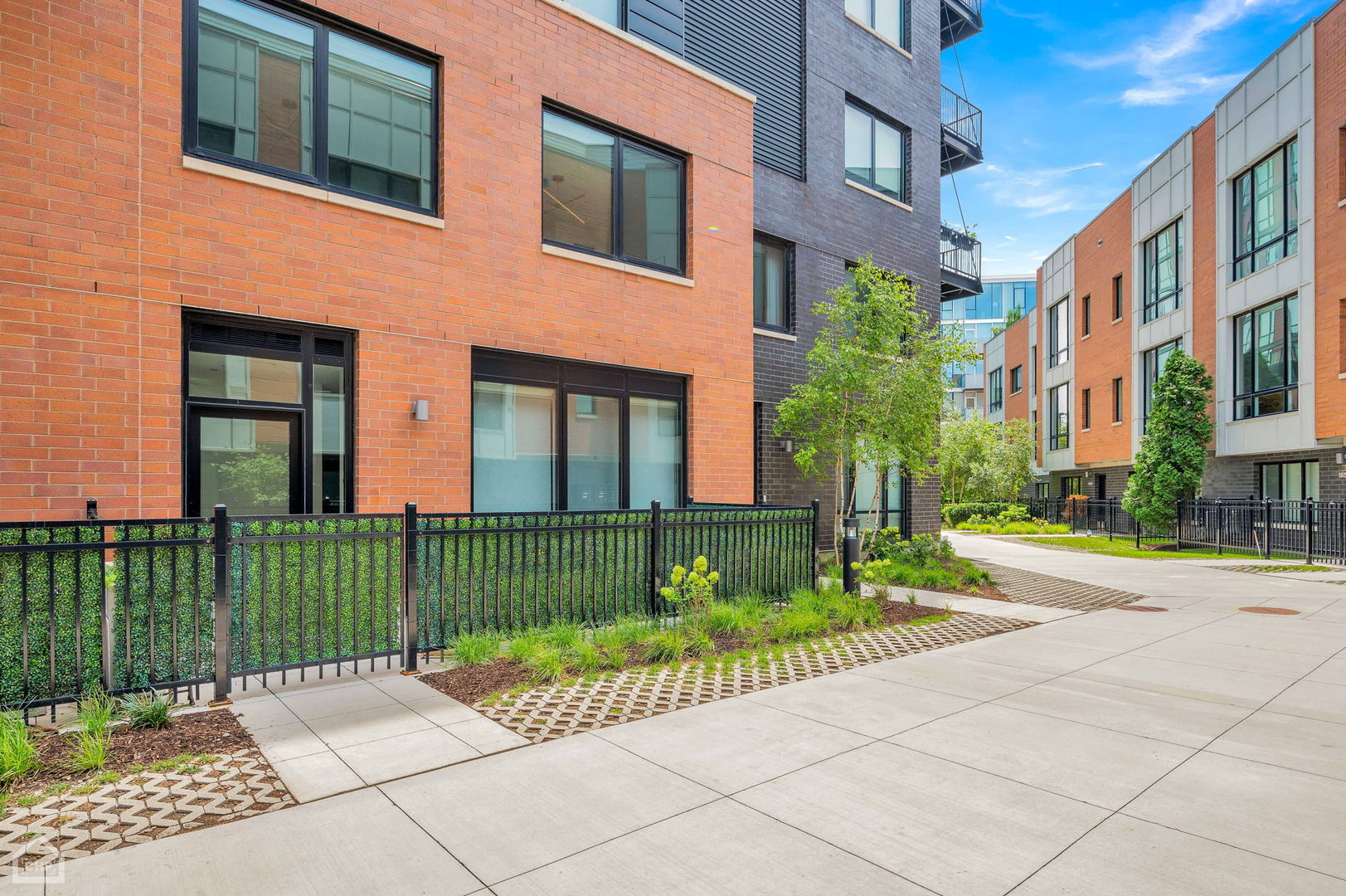 Modern brick residential buildings beside a landscaped courtyard walkway and black metal fencing.