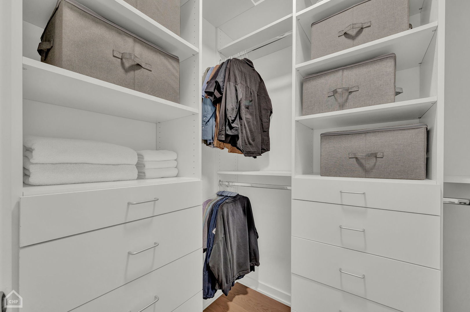 Walk-in closet with white built-in drawers, gray storage bins, hanging clothes, and folded towels.