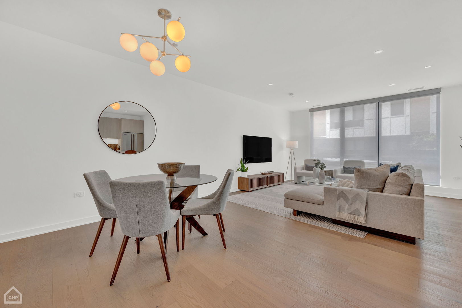 Open-plan living and dining room with a beige sectional, round glass dining table, and large shaded windows.