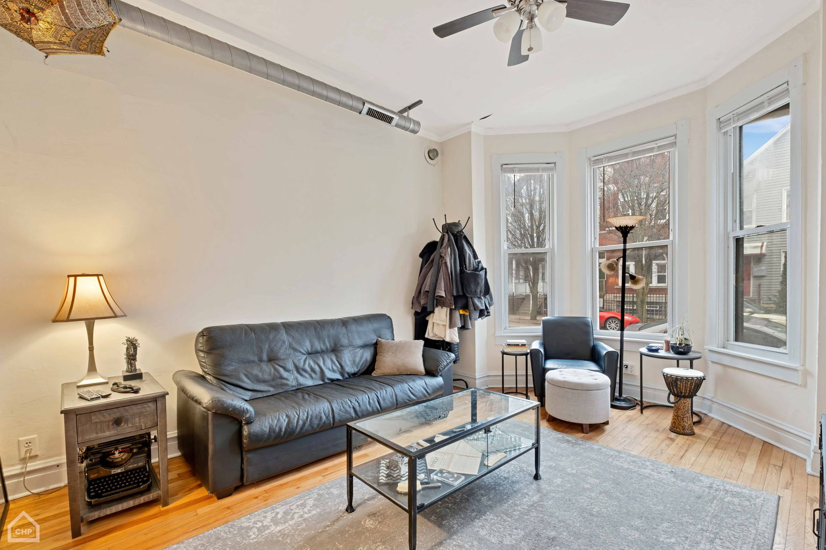 Living room with a gray leather sofa, glass coffee table, and three tall street-facing windows.