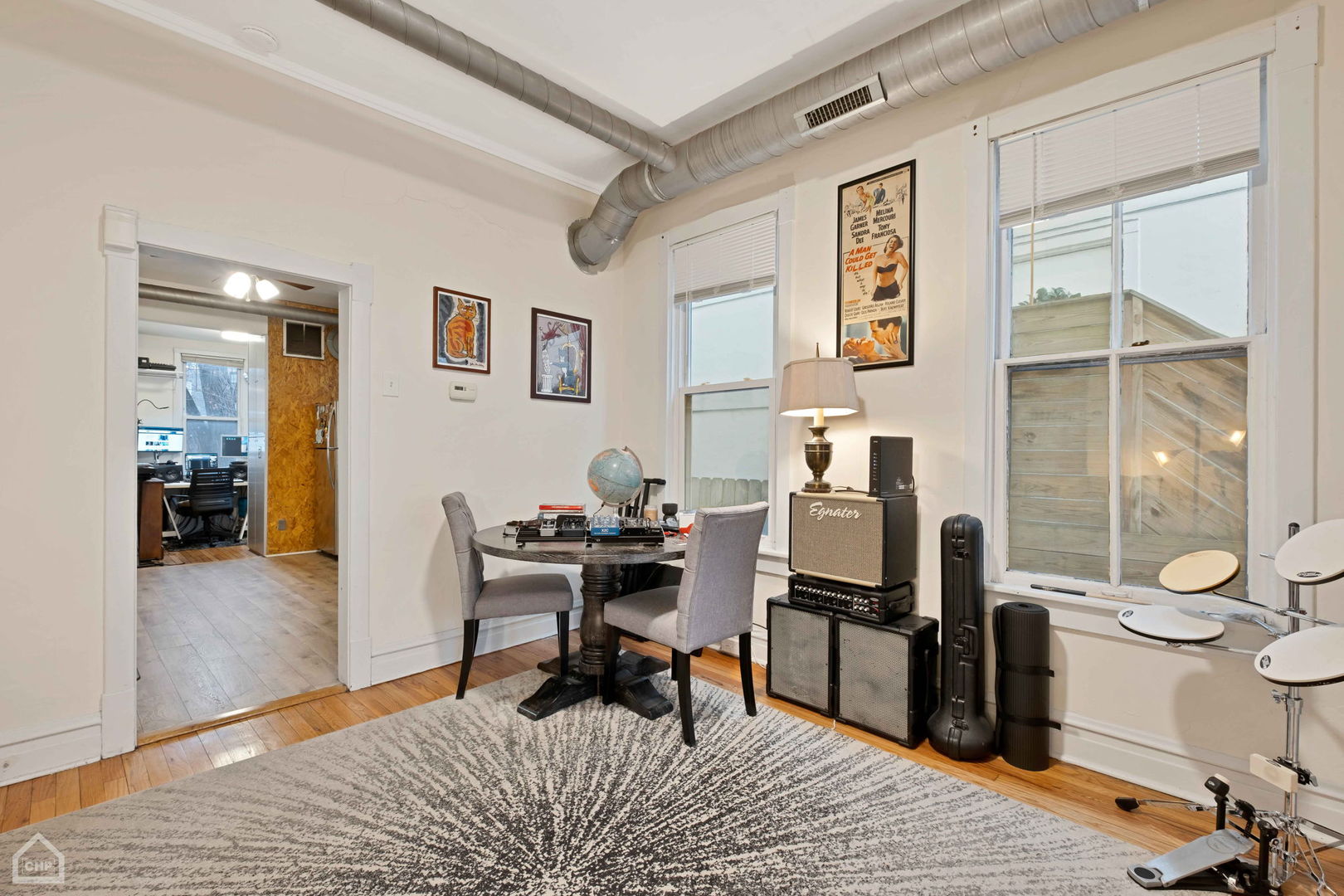 Furnished room with a round table, guitar amplifiers, and an electronic drum kit beside large windows.