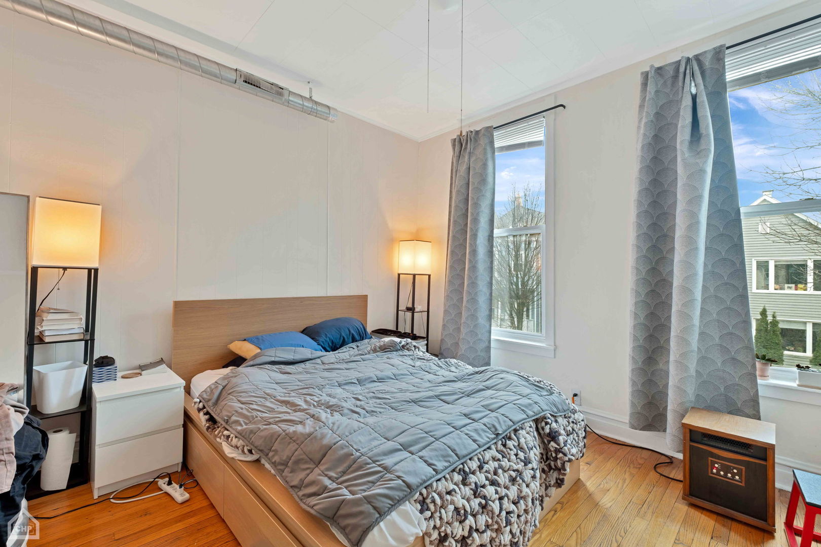 Bedroom with a wooden bed, gray quilt, hardwood floors, and tall curtained windows.