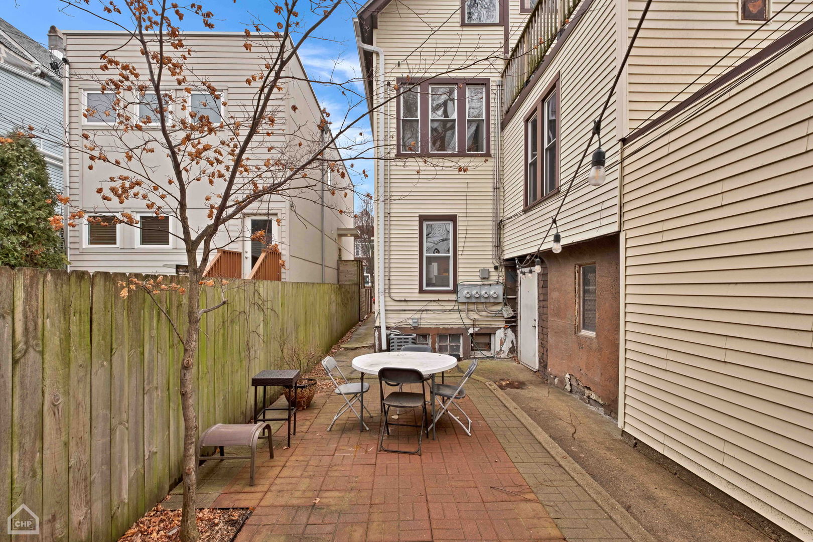 Brick patio with a round table and chairs beside a beige-sided home and wooden fence.