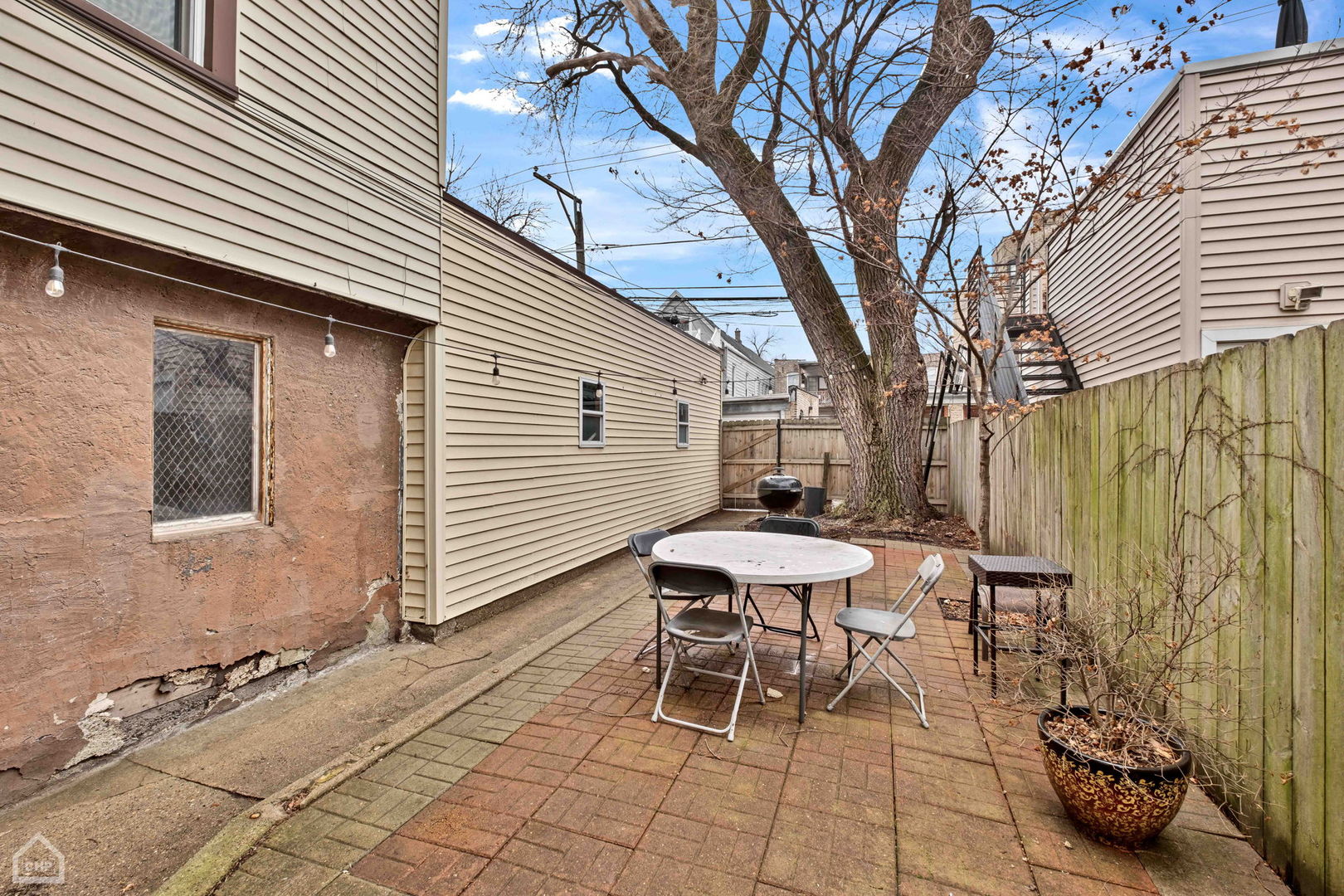 Backyard patio with a round table, folding chairs, wooden fence, and large tree.