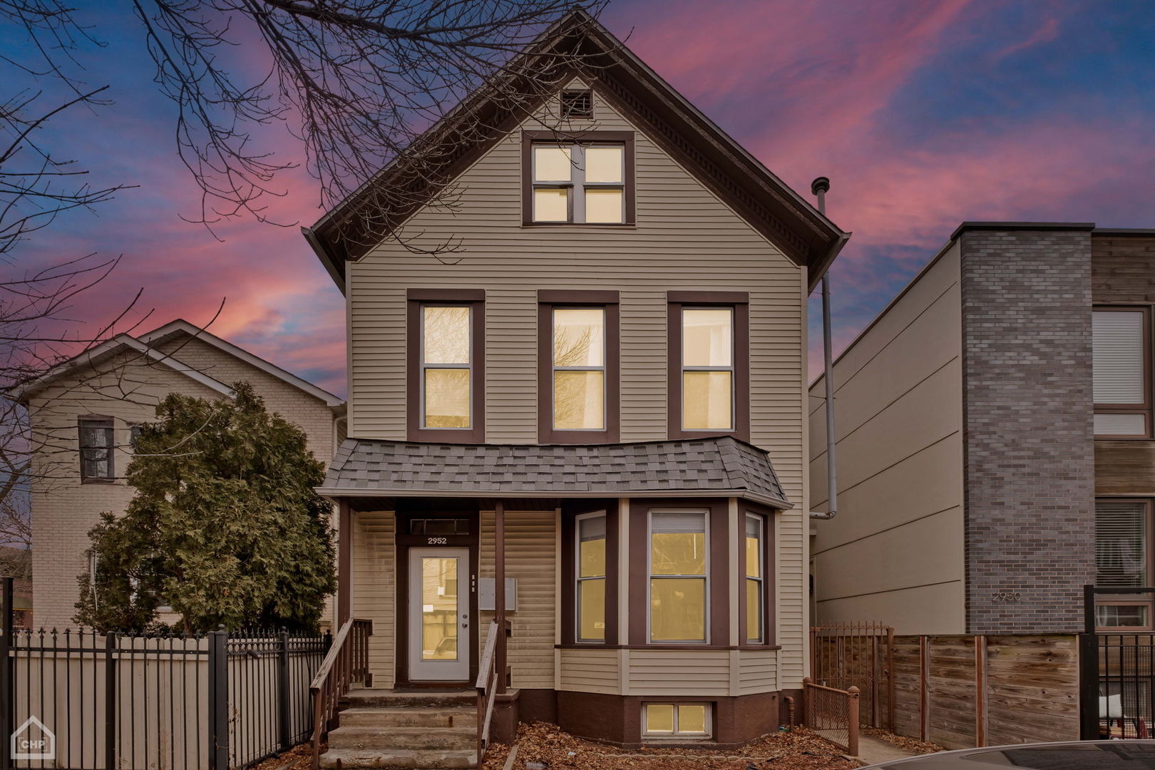 Beige gabled house with a bay window and illuminated windows beneath a pink dusk sky.