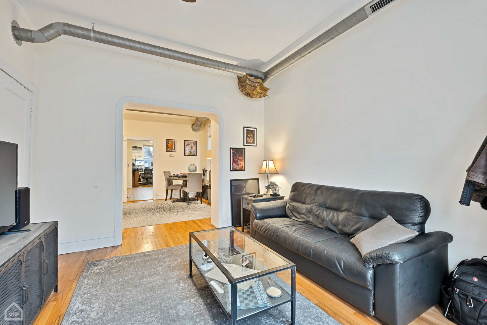 Living room with a black leather sofa, glass coffee table, hardwood floors, and exposed ceiling ductwork.
