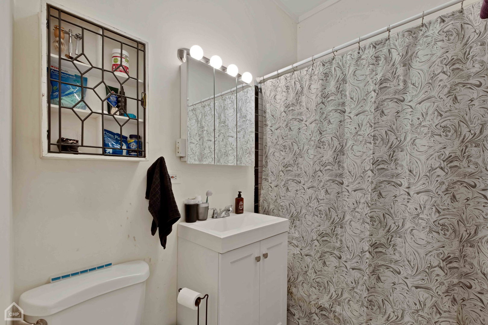Bathroom with a white vanity, mirrored cabinet, toilet, and patterned gray shower curtain.