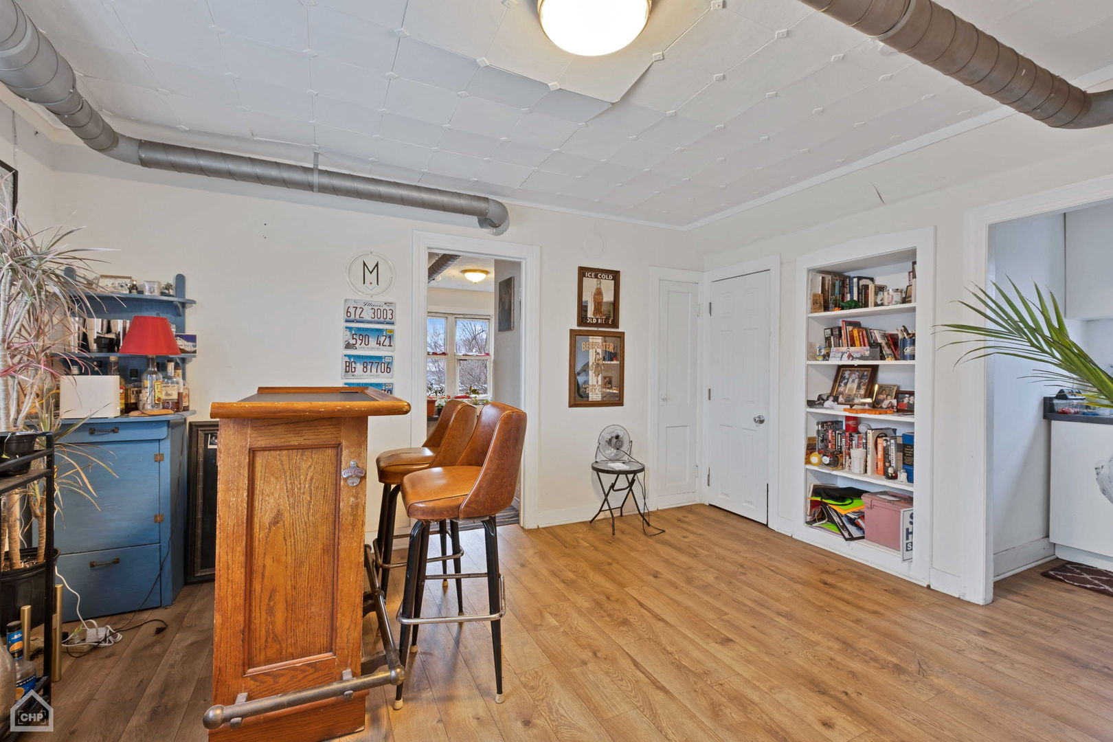 Furnished bar area with a wooden counter, leather stools, built-in bookshelves, and exposed ceiling ducts.