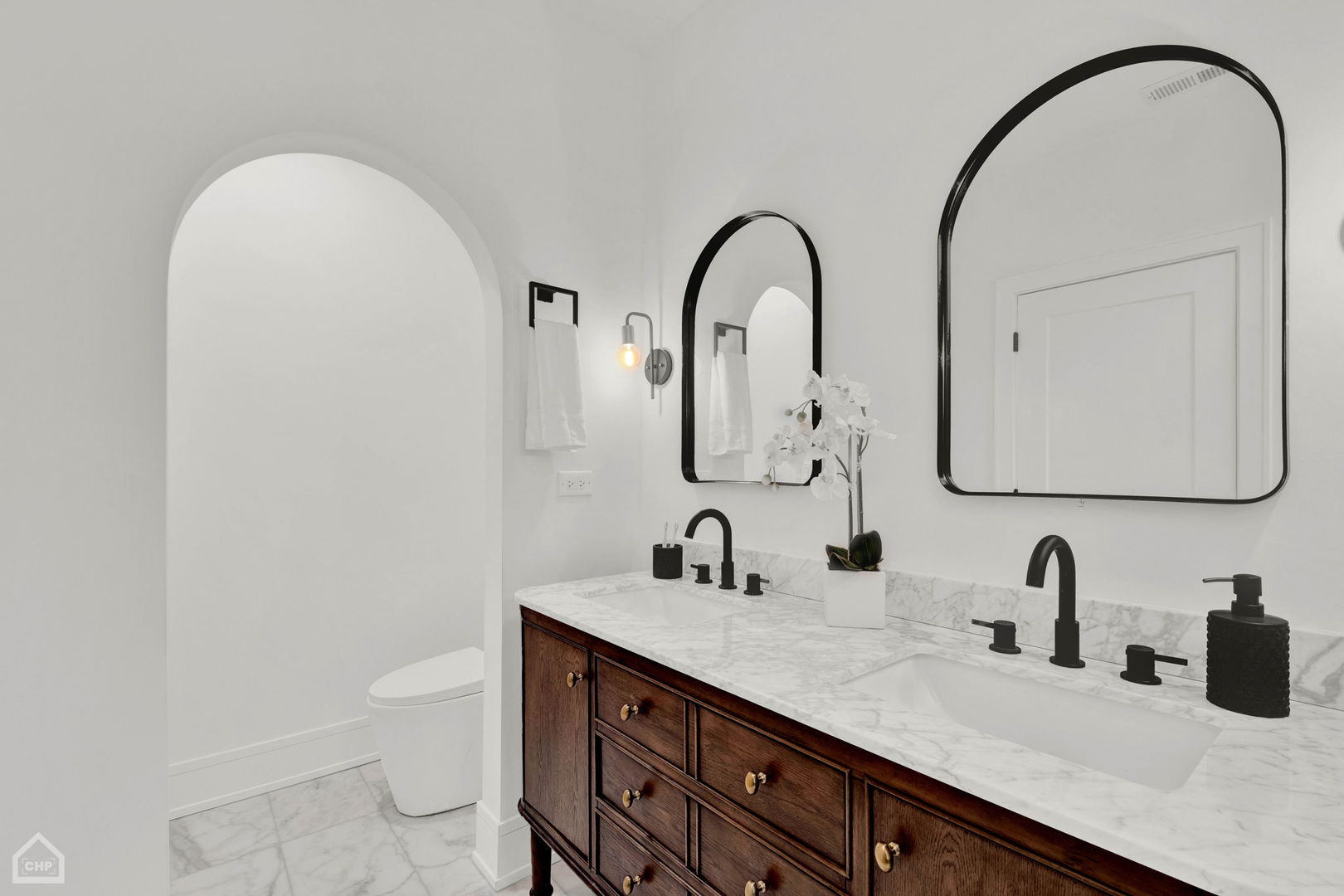 Bathroom with a double-sink dark wood vanity, marble surfaces, black arched mirrors, and a separate toilet alcove.