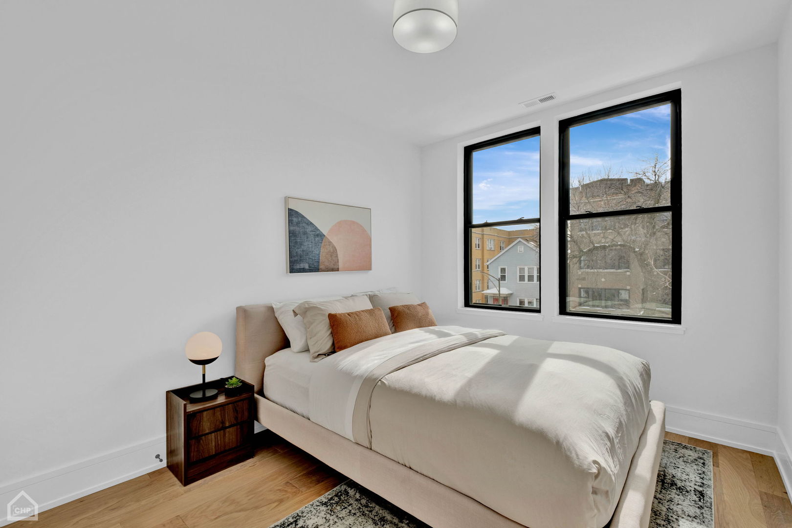 Staged bedroom with a cream bed, rust-colored pillows, and two large black-framed windows.